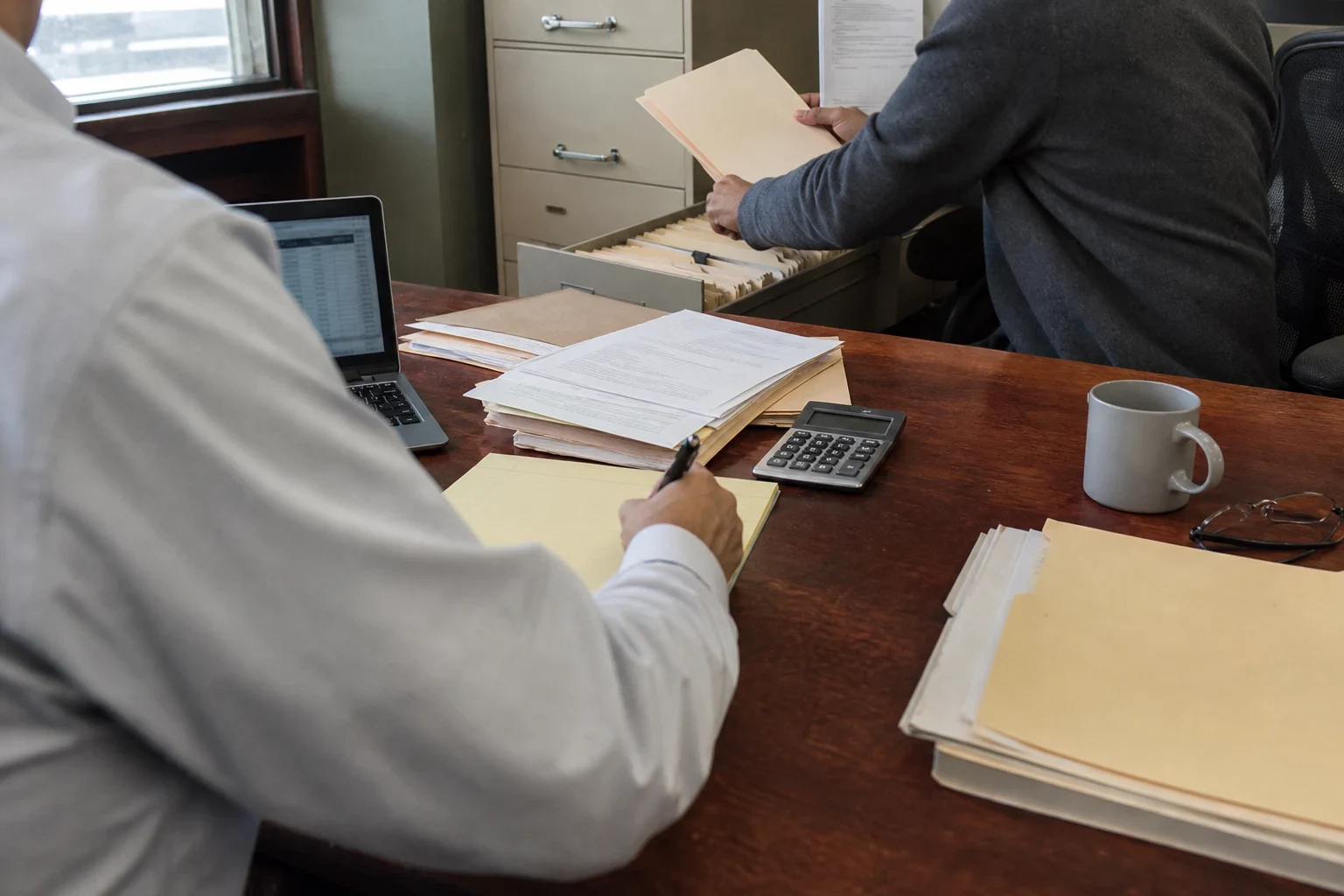Agent in a light dress shirt takes notes on a yellow legal pad at a wooden desk while a client in a gray sweater pulls a file from an open filing cabinet