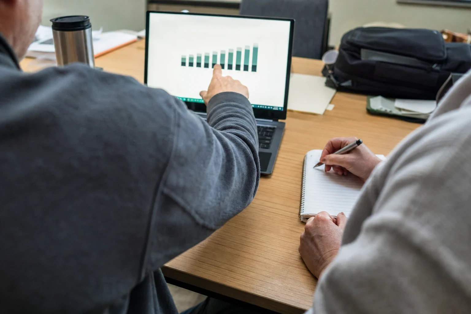 Over-the-shoulder view of a man pointing at a bar chart on a laptop at an office desk while another person takes notes in a spiral notebook