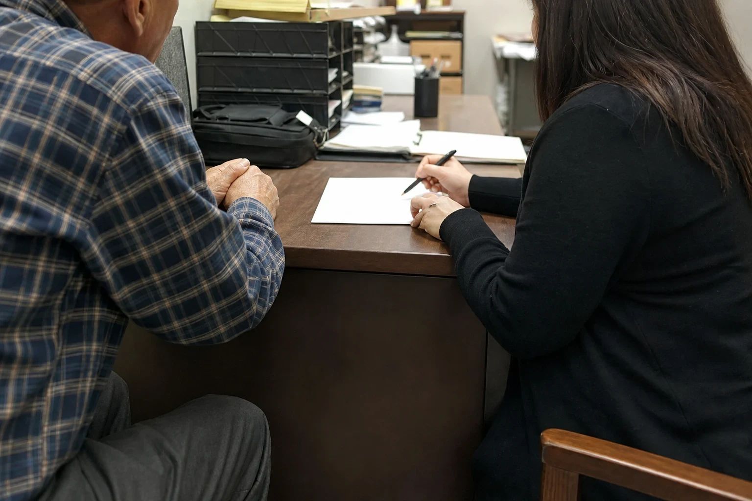 An older man in a plaid shirt sits at a wooden office desk beside a woman in black who holds a pen over a sheet of paper, with file trays and a bag behind them